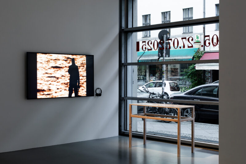 A video screen showing a silhouetted figure at sunset is mounted near a street-facing gallery window, accompanied by headphones and a bare wooden table.