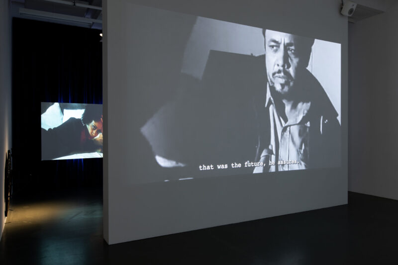 Two video projections face each other in a darkened gallery space, one showing a black-and-white film with subtitles, the other a color image of a woman on a bed.