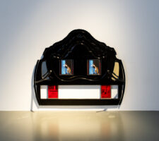 A black ornate structure resembling a headboard incorporates embedded screens and red panels, dramatically lit against a gray floor.