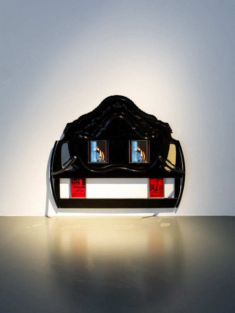 A black ornate structure resembling a headboard incorporates embedded screens and red panels, dramatically lit against a gray floor.