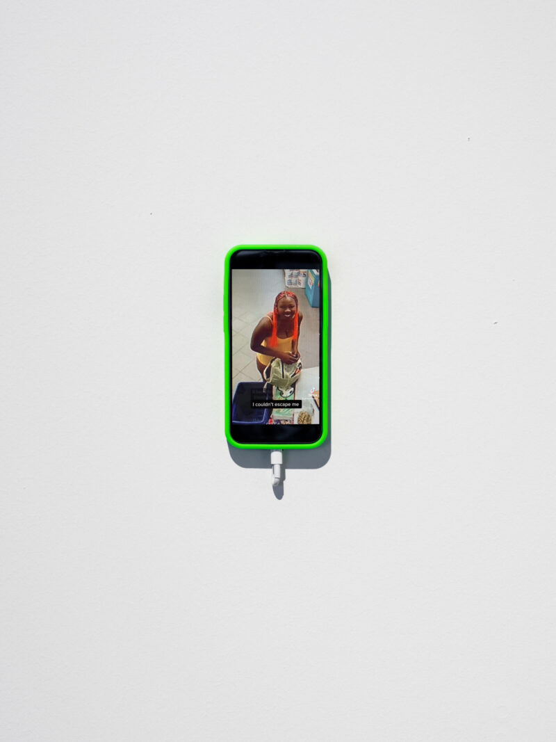 A green-cased smartphone mounted on a white wall displays a vertically split screen with a woman on a video call and the caption “I couldn’t escape me.”