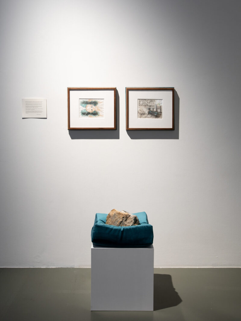 Two framed drawings hang above a teal cushion holding a rock, all mounted on a white pedestal in a minimal display.