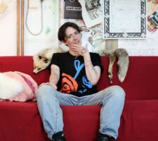 Artist Elif Saydam sits on their red couch in their studio