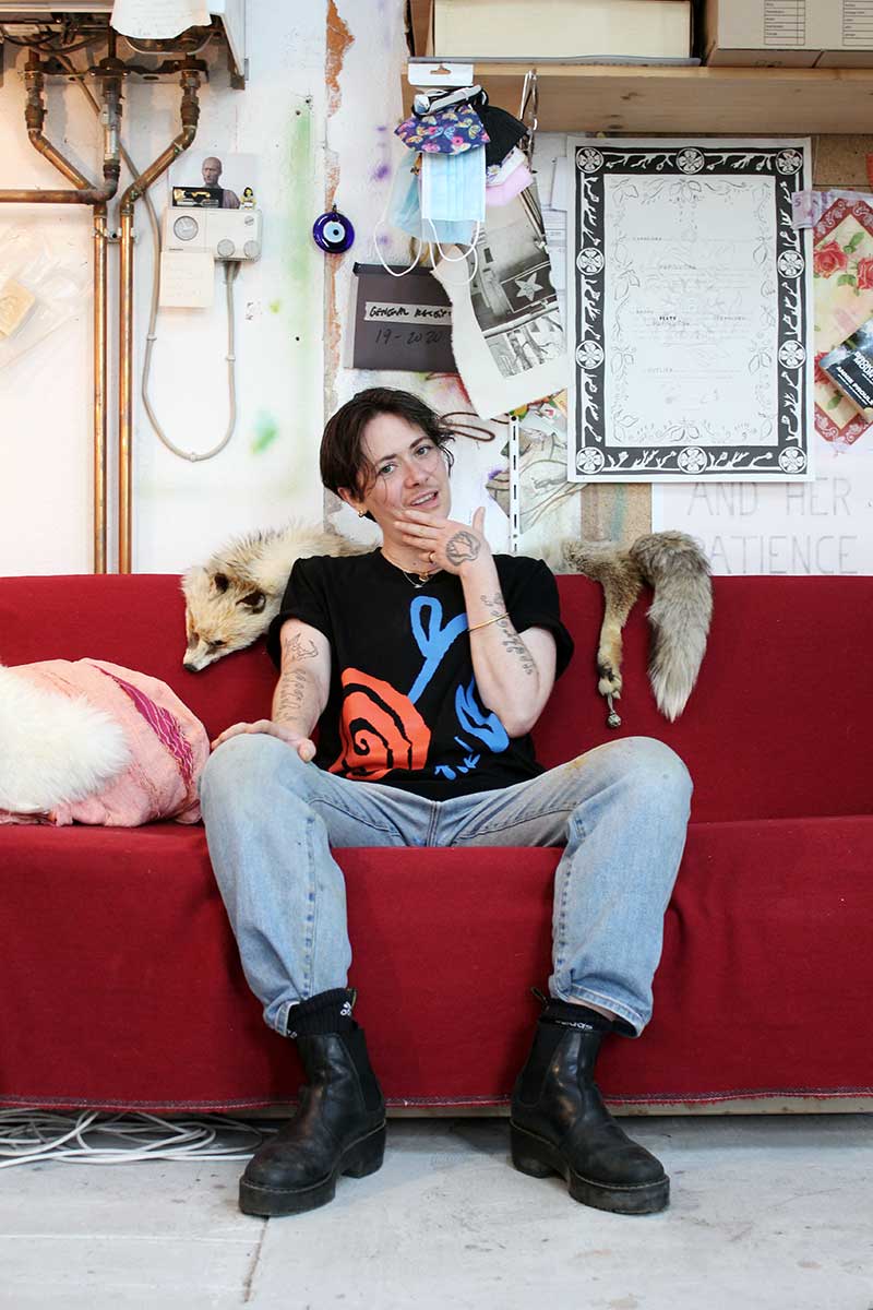 Artist Elif Saydam sits on their red couch in their studio