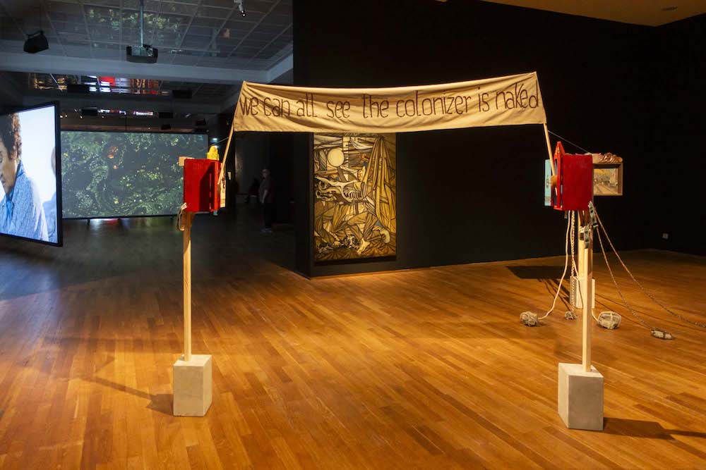 art installation with banner saying "we can all see the colonizer is naked"