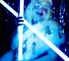 A performe of Drag Syndrome is holding neon light tubes, wearing a metal dress and a white wig