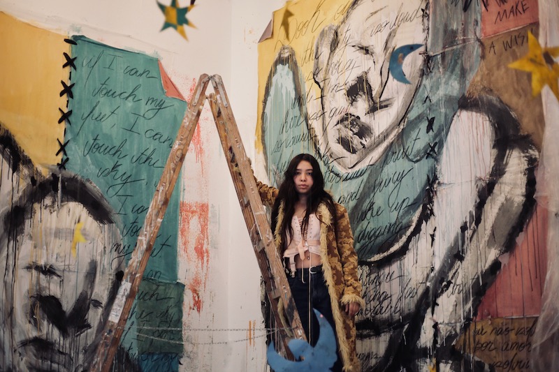 artist with long dark hair stands on a ladder in front of two large scale paintings of figures outlined in black and surrounded my pink, yellow and turqoise color blocks