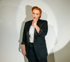 a person with short, sleek orange-colored hair stands in front of a circular stage light, wearing a black lounge suit and white shirt, she looks into the camera