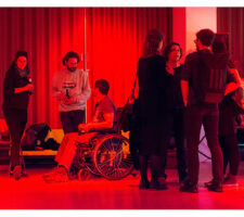 in a red-lit room with a curtain against the back wall, a group of people stand or sit in wheelchairs, speaking to each other and having a drink