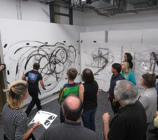 participants in a workshop for partially sighted and blind people in architecture pictured in front of two large wall canvases, with the teacher drawing black circular figures on them