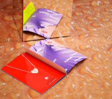 a close up of the open book with a heart shaped locket on a red page, the book is sitting next to a mirrored plinth and on a floor covering that looks like water droplets