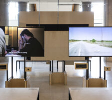 a photo of an exhibition installed in HKW with several screens and desks with chairs
