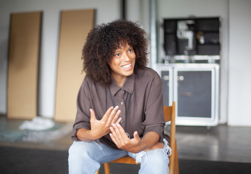 the artist isabel lewis sits on a wooden chair in her studio, looking off to the side and mid conversation, wearing a grey brown button down shirt and blue jeans ripped at the knees