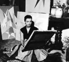 A black and white photographer of the artist Lygia Clark sitting in her studio amidst her geometric paintings, from 1950