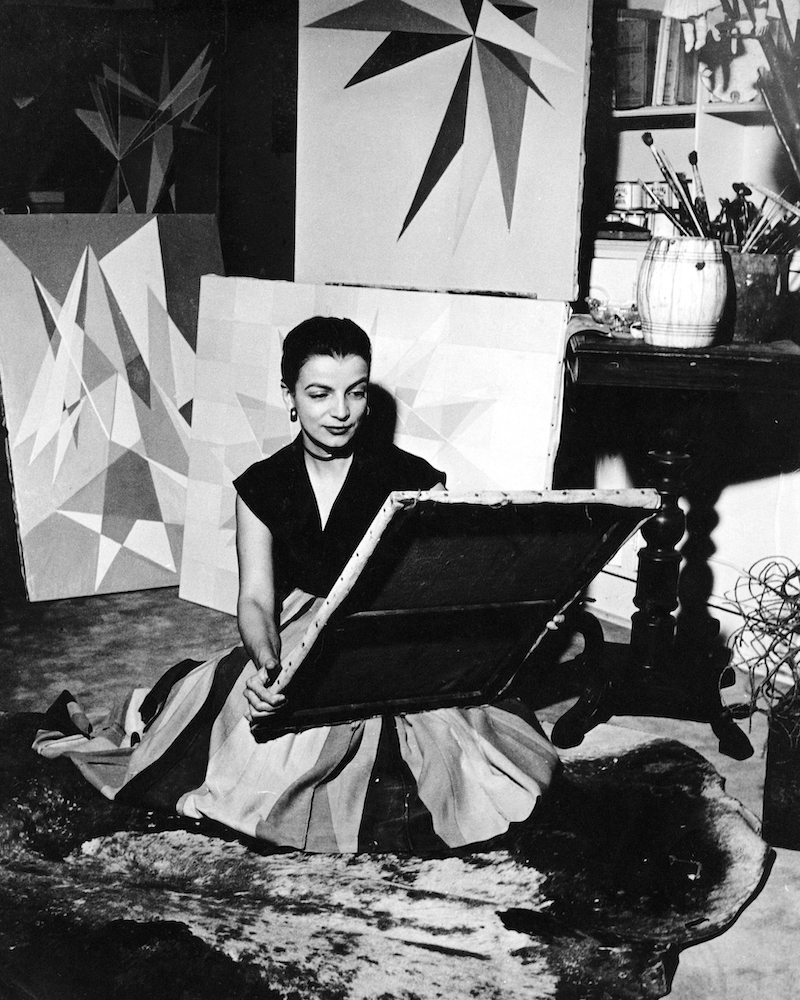 A black and white photographer of the artist Lygia Clark sitting in her studio amidst her geometric paintings, from 1950