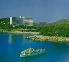 a grainy postcard picture in blue and green on an island with a ferry boat in the foreground