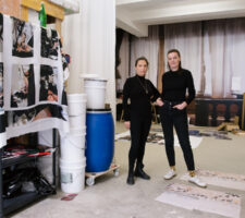 two artists stand in their studio space, next to shelves full of materials, both dressed in black