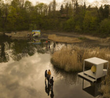 a drone aerial photo of the water basin, with art installations spread across the water and people in rain boots looking at them