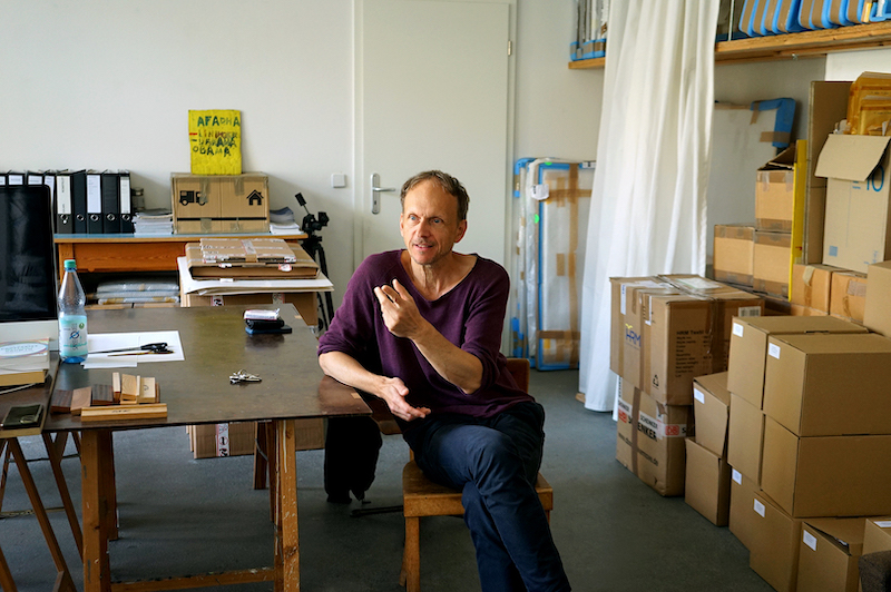 Julian Rosefeldt sits at the desk in his studio, his hand gesticulating while he speaks