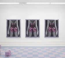 three identical portraits of a woman in a sequined dress are mounted on a gallery wall, with a checkered floor in the foreground and some undefined mauve box sculptures on the floor